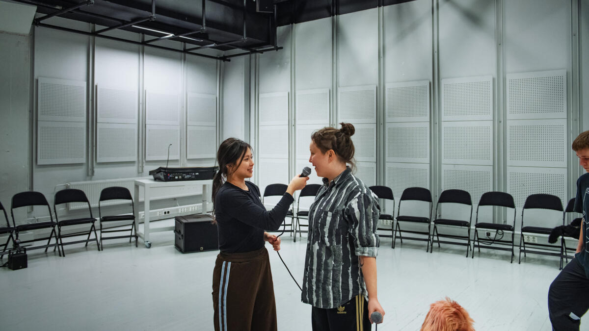 Kaksi opiskelijaa seisovat vaaleassa studiosalissa. Vasemmanpuoleinen opiskelija pitää mikrofonia kädessään haastatellakseen vierustoveriaan.