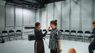 Kaksi opiskelijaa seisovat vaaleassa studiosalissa. Vasemmanpuoleinen opiskelija pitää mikrofonia kädessään haastatellakseen vierustoveriaan.