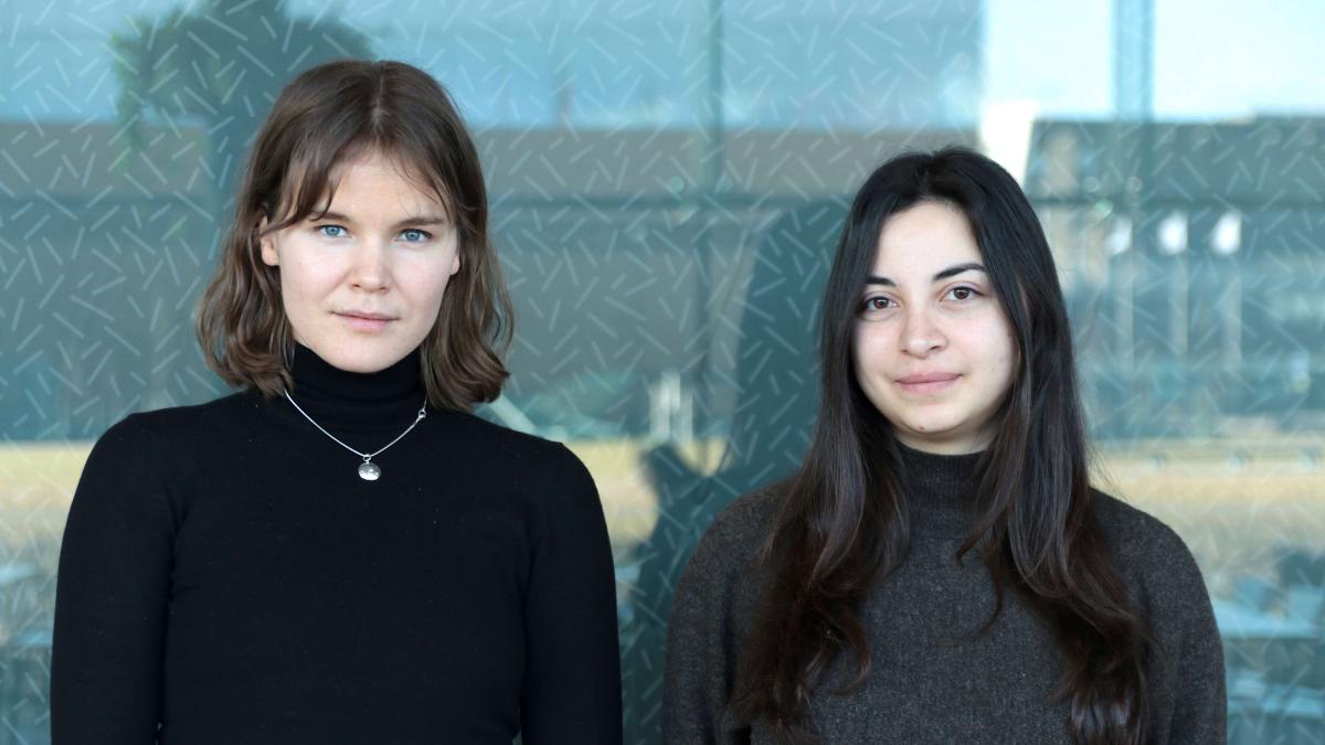 Kaksi naista seisoo vierekkäin lasiseinän edessä ja katsoo suoraan kameraan.