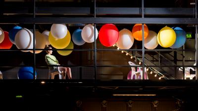 Actor sitting in a chair at a performance stage setting, large colourful balloons above their head.