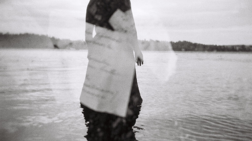 A black and white image of a figure wearing a long black skirt, overlaid with double-exposed blurry text on white paper.