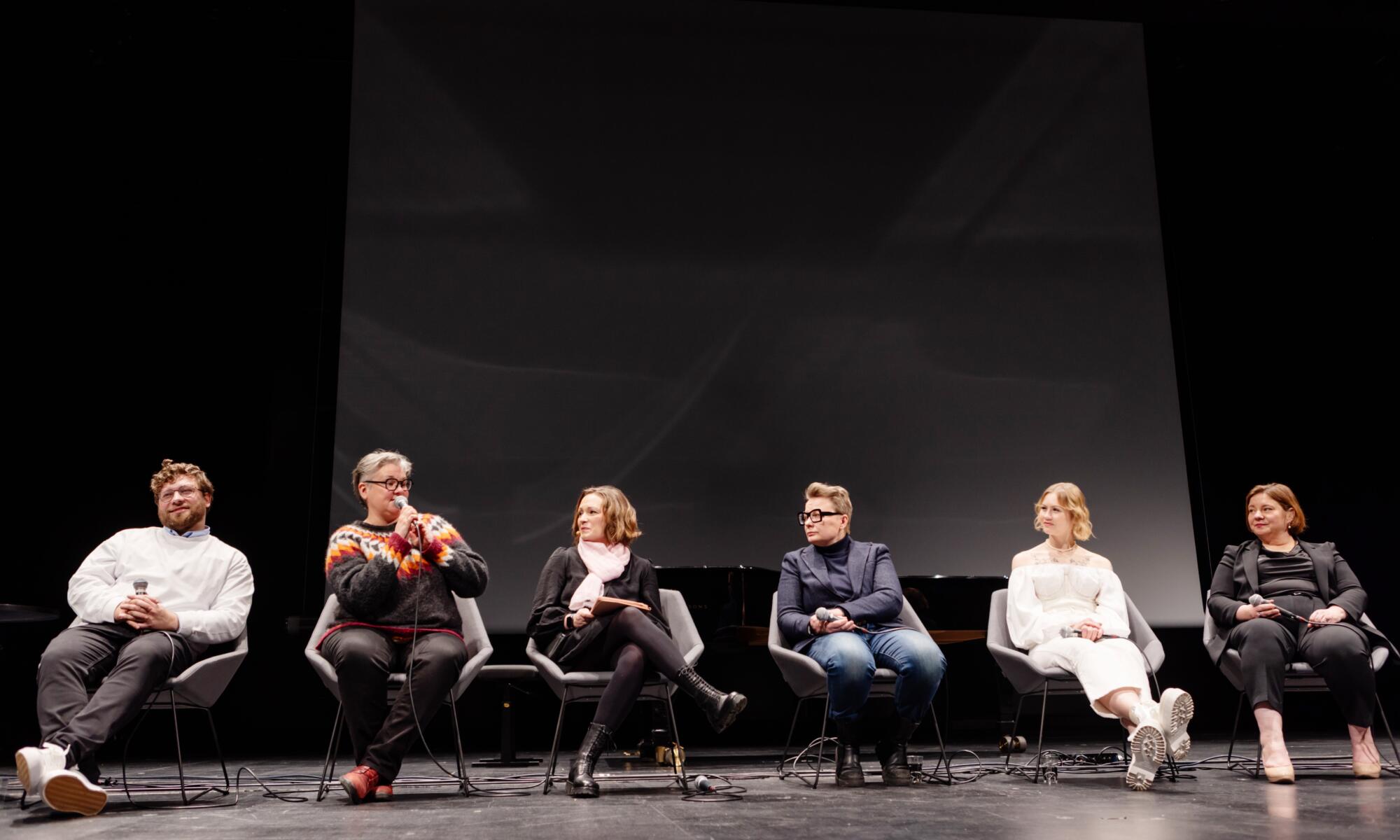 People sitting and discussing in a row on a stage with black background.