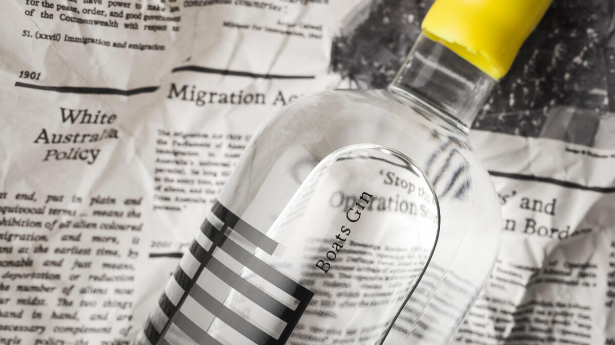 A closeup of a clear bottle with the text "boat's gin" on it, agains crumpled newspaper.