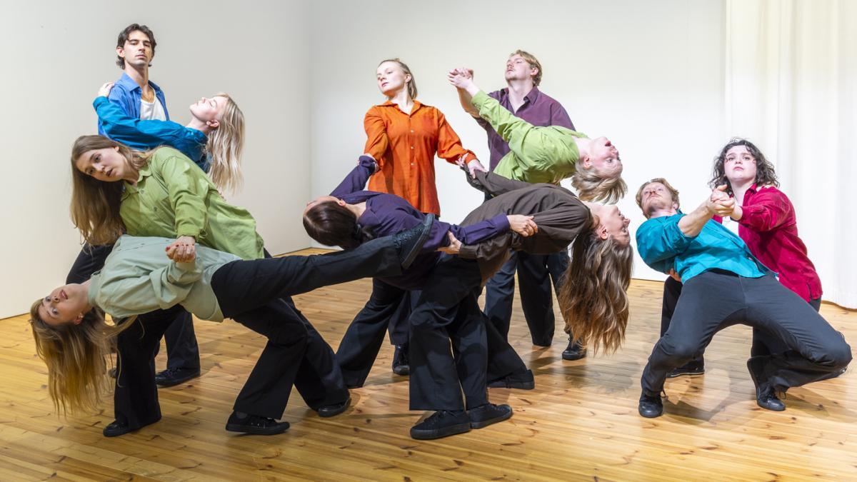 Kuvassa 11 ihmistä liikuntasalissa, jossa puulattia. Ihmiset ovat eri asennoissa.