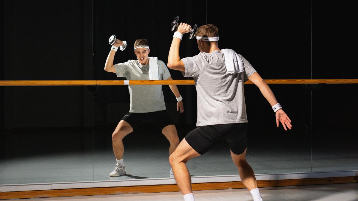 Kuvassa henkilö seln kameraan peilin edessä. Hänellä on käsipaino vasemmassa kädessä, pyyhe olalla, hikinauha päässä ja ranteessa.