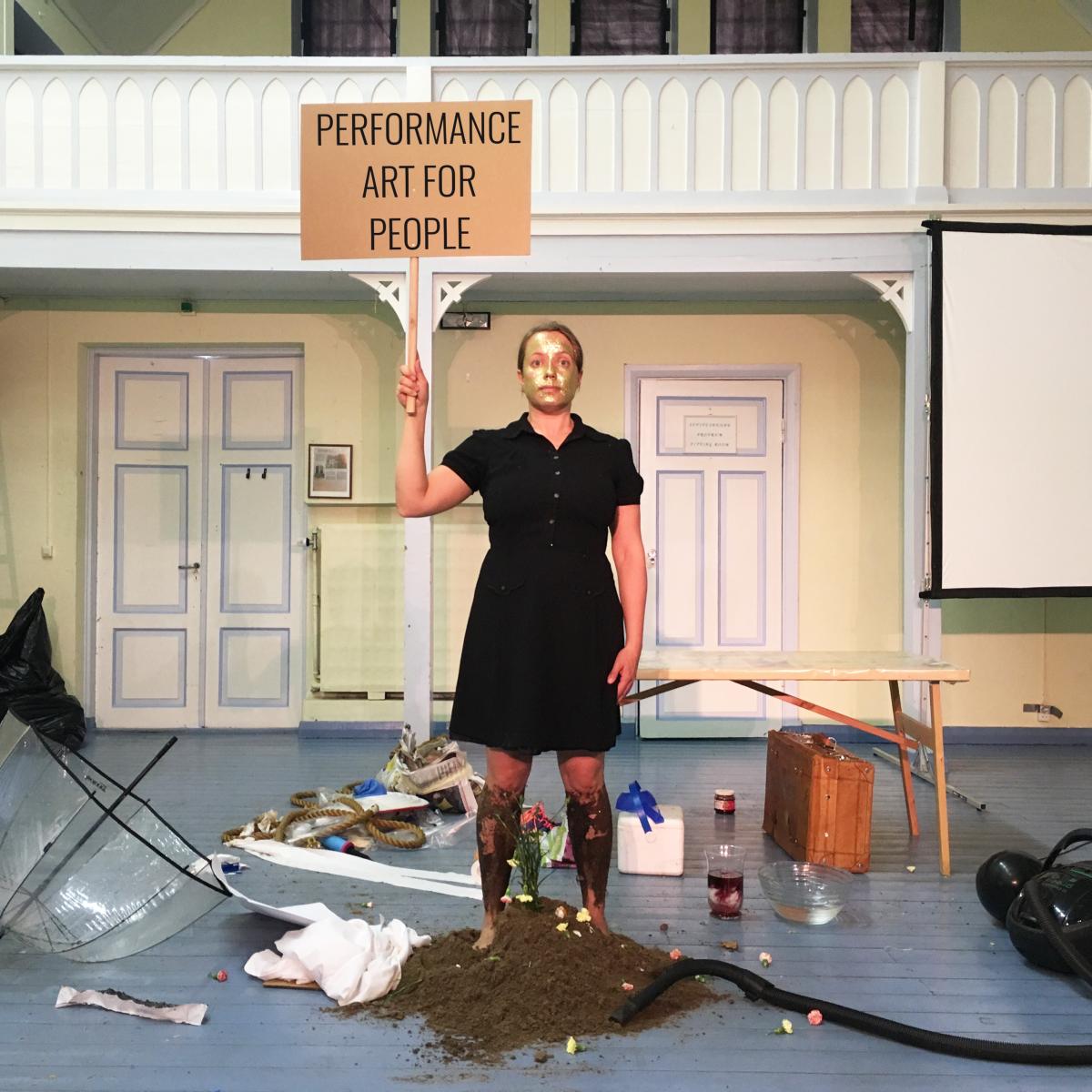 Leena Kela is standing in the middle of a room holding a sign that says 'Performance art for people'. There are objects scattered around her and she is standing on a pile of soil and she has golden paint on her face.
