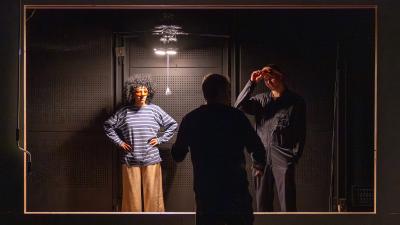 Ohjaaja keskustelee roolivaatteisiin pukeutuneiden näyttelijöiden kanssa mustassa tilassa.