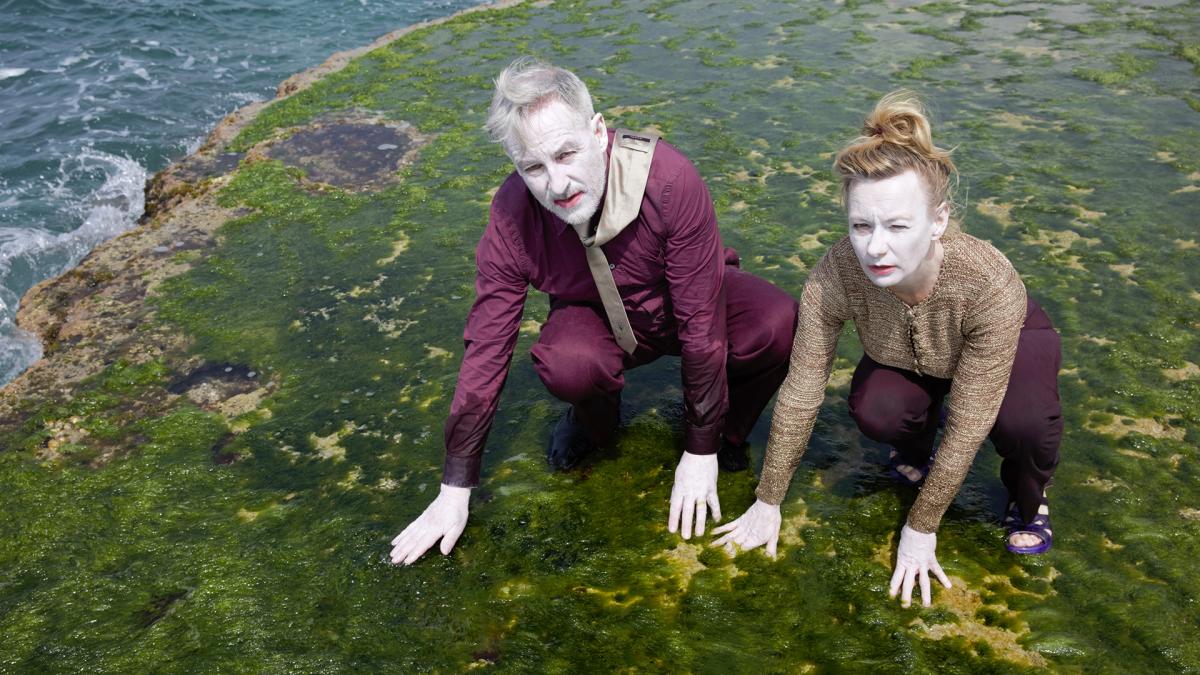 Taiteilijat Simon Häggblom ja Karin Lind valkoisella kasvomaalilla maalattuina katsovat kameraan, ovat kyykyssä ja koskevat maata joka on vihreää levää.