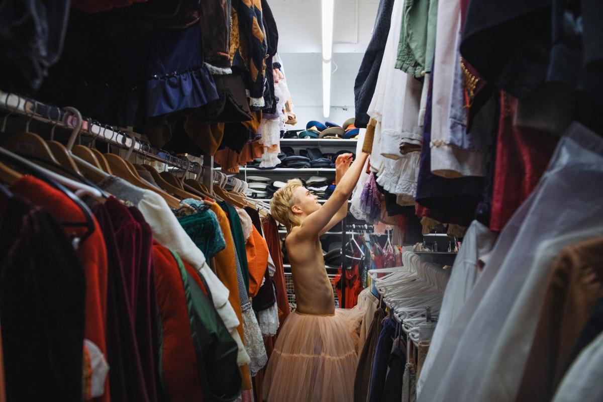 Opiskelija kurottaa kohti vaatetta puvustamon vaaterekkien välissä. Hänellä on yllään vaaleanpunainen alushame.