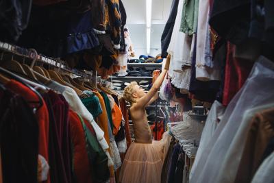 Opiskelija kurottaa kohti vaatetta puvustamon vaaterekkien välissä. Hänellä on yllään vaaleanpunainen alushame.