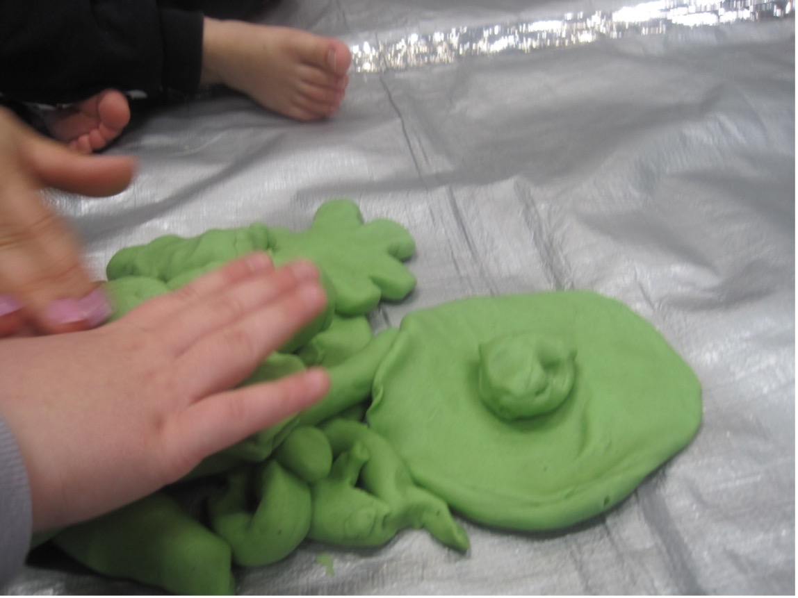 Valokuva, jossa lapset muovailevat vihreästä muovailutaikinasta erilaisia muotoja hopeanvärisellä alustalla. Kuvassa näkyy useita käsiä sekä taikinasta tehtyjä pyöreitä, pitkulaisia, kukkamaisia ja litteitä muotoja, taustalla paljaat jalat.