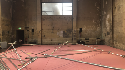 Tyhjä teollisuushalli, jossa on korkea katto ja kuluneet betoniseinät, joissa näkyy lohkeilevaa rappausta ja ikää. Keskellä kuvaa vaaleanpunaisella matolla makaa sikin sokin rakennustelineen metallisia osia. Luonnonvalo pääsee sisään takaseinän isosta ikkunasta. Takaseinän vasemmalla reunalla on ovi, jonka yläpuolella on exit-valo.