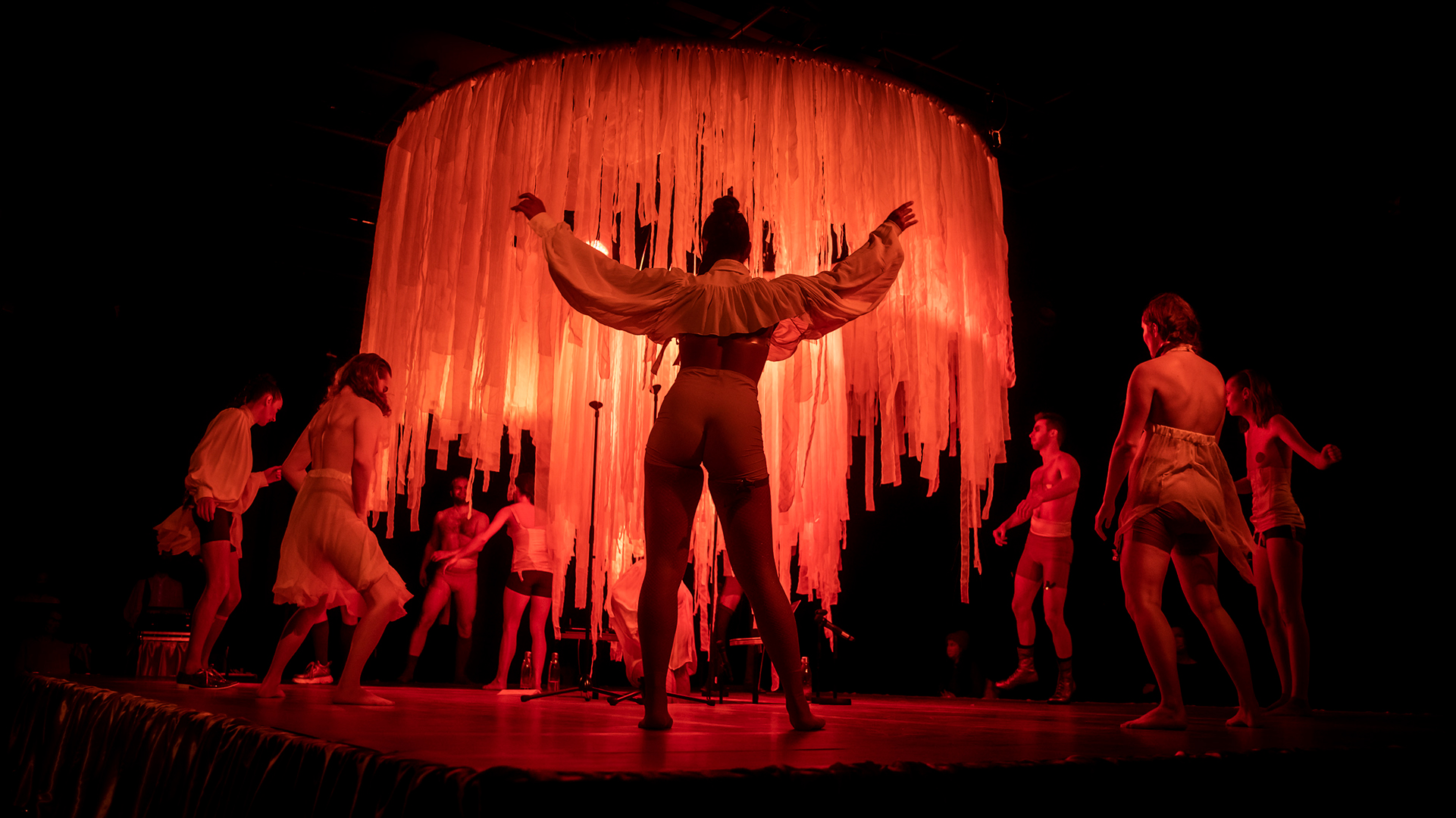 Performers on stage with backs facing the camera. Dark background and reddish lighting, large chandelier in the middle of the stage.