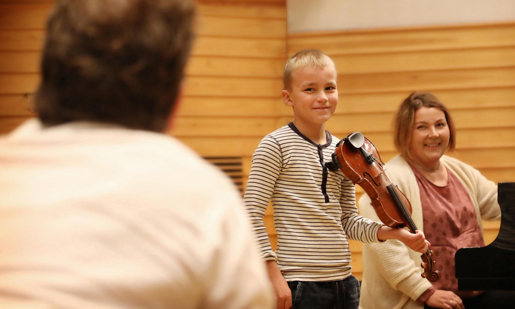 Nuori viulistipoika ja pianisti katsovat hymyillen kameraan