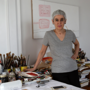 Taiteilija Mira Schor seisoo työpöytänsä vieressä. Pöydällä on taidemaalarin työvälineitä.