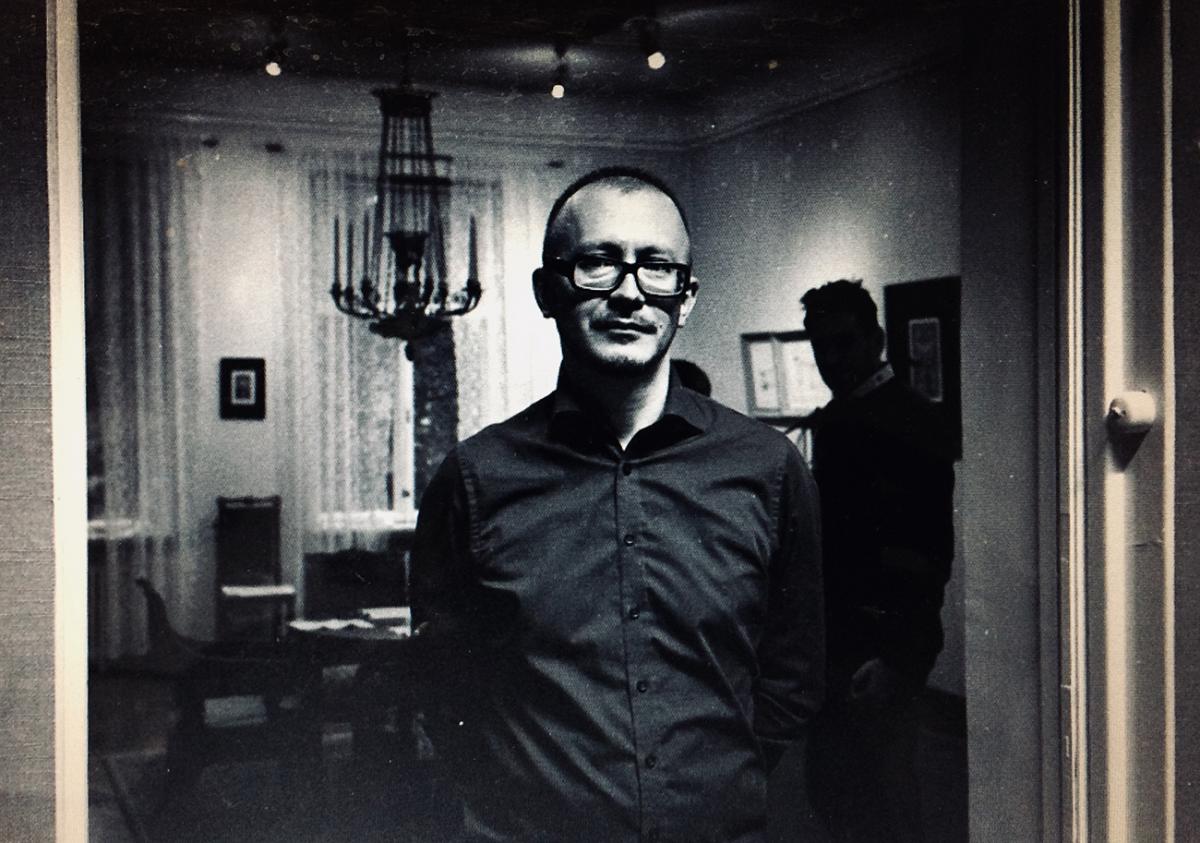 Ilya Orlov katsoo kameraan mustavalkoisessa kuvassa. Kuvan taustalla on huone, jossa ihmisiä ja suuri kynttiläkruunu katossa.
