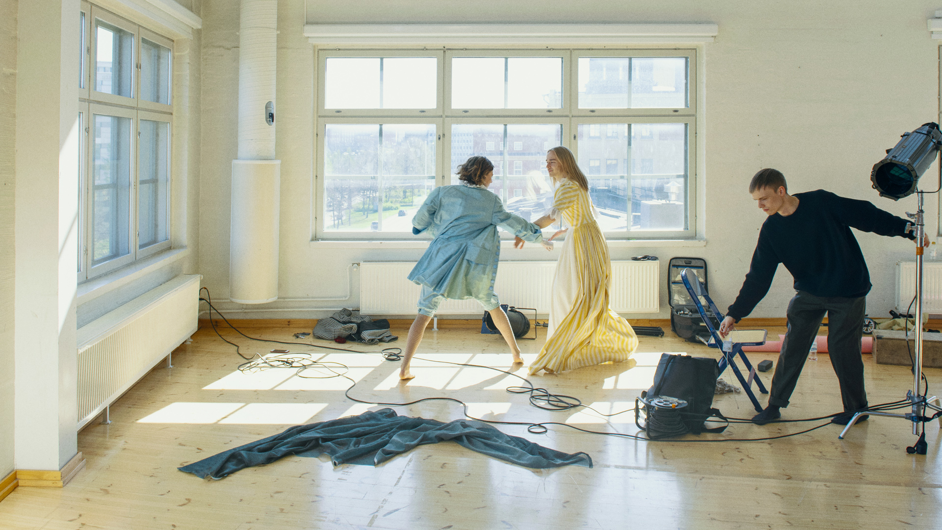 Three persons in a bright and spacious rehearsal room. Two are wearing costumes, third person is holding wires of lighting equipment.