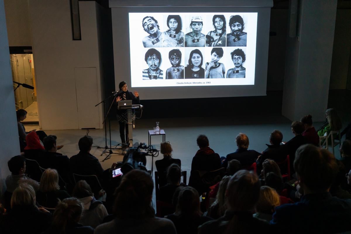 Andre Giunta luennoi yleisön edessä. Puhujan takana on heijastettu valokuvateos, jossa on ihmisiä.