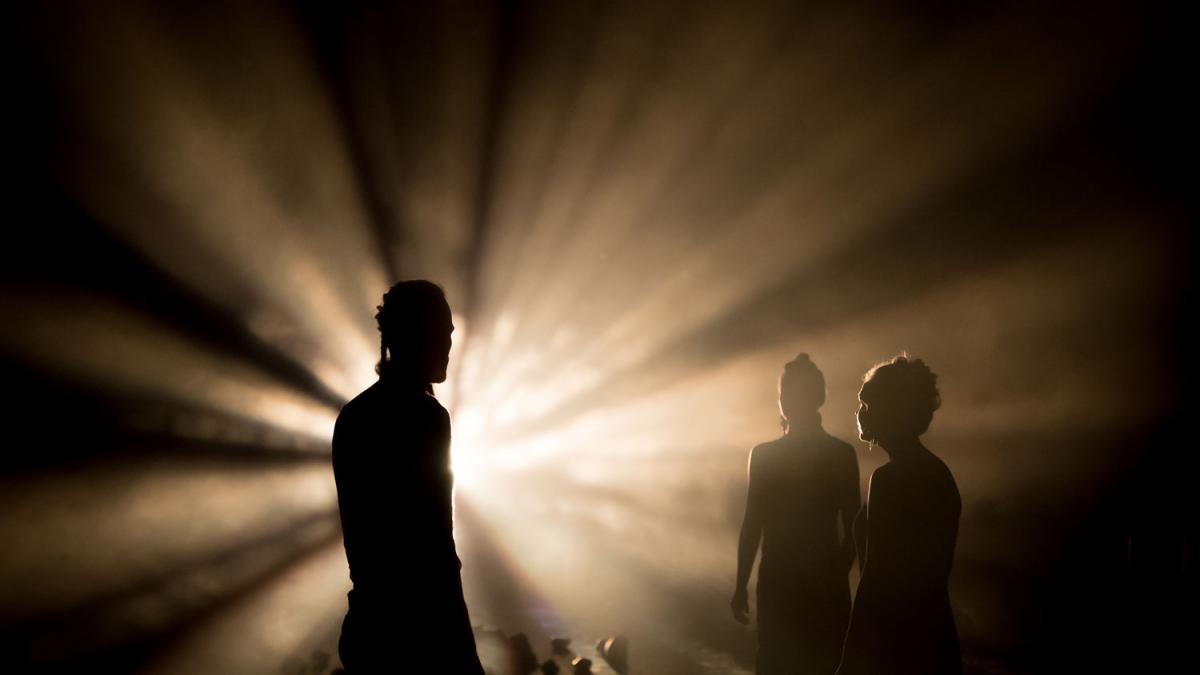 Kolme hahmoa teatterilavalla, takaa loistaa valo.
