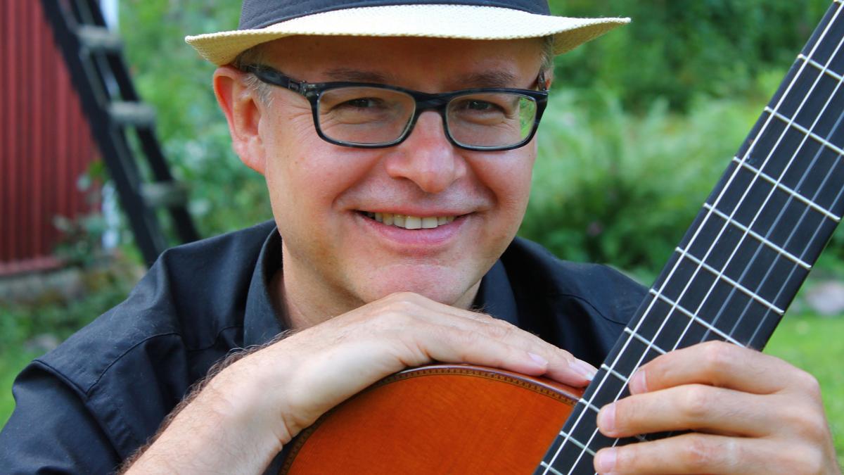 Andrzej Wilkus sitter och lutar sig på en gitarr och ler mot kameran.