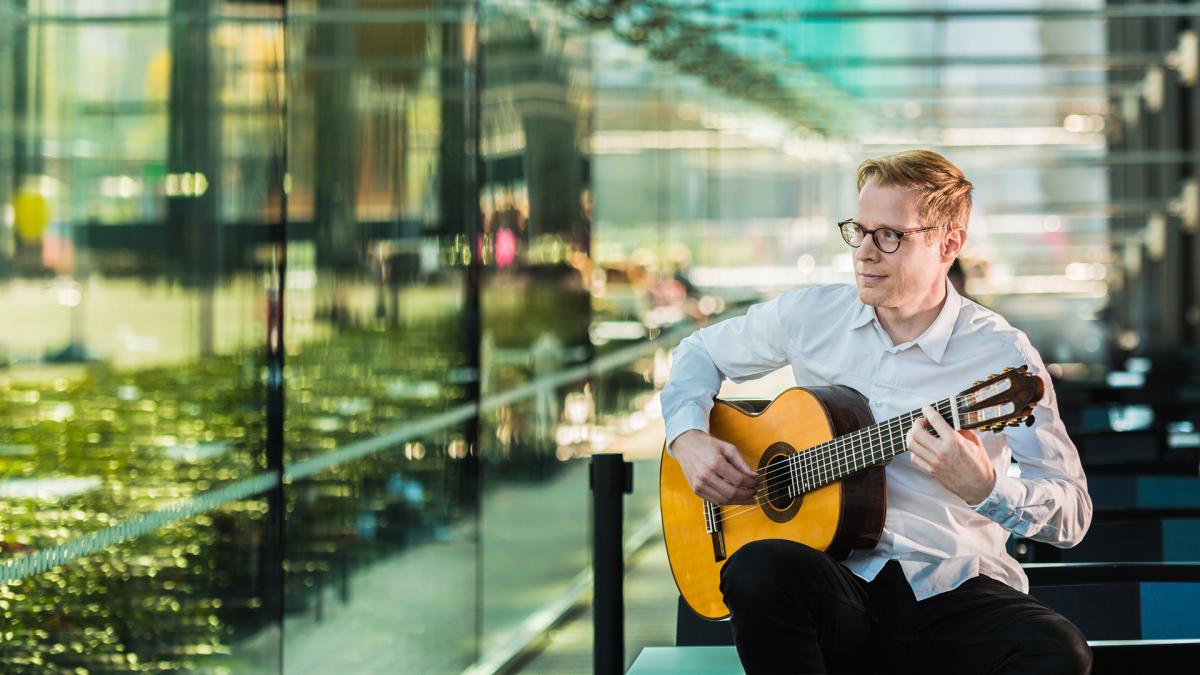 Janne Malinen sitter framför fönsterväggen i Musiikkitalo-kaféet och spelar gitarr. Han ser ut och ler.