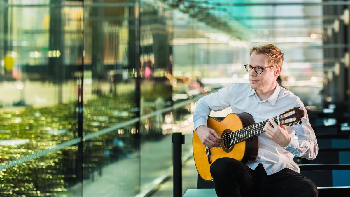 Janne Malinen sits in front of the window wall in the Musiikkitalo café and plays guitar. He looks out and smiles.