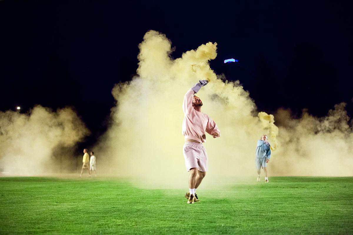 Ihmisiä urheiluasuissa jalkapallokentällä keltaisen savun keskellä.