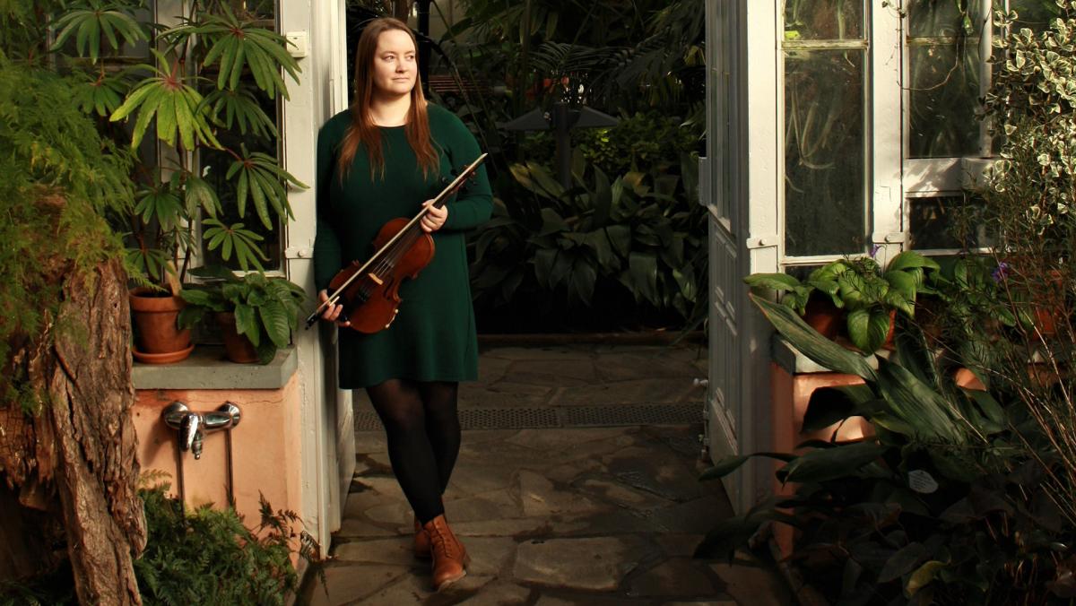 Maisa Saari poseeraa viulun kanssa viherkasvien seassa.