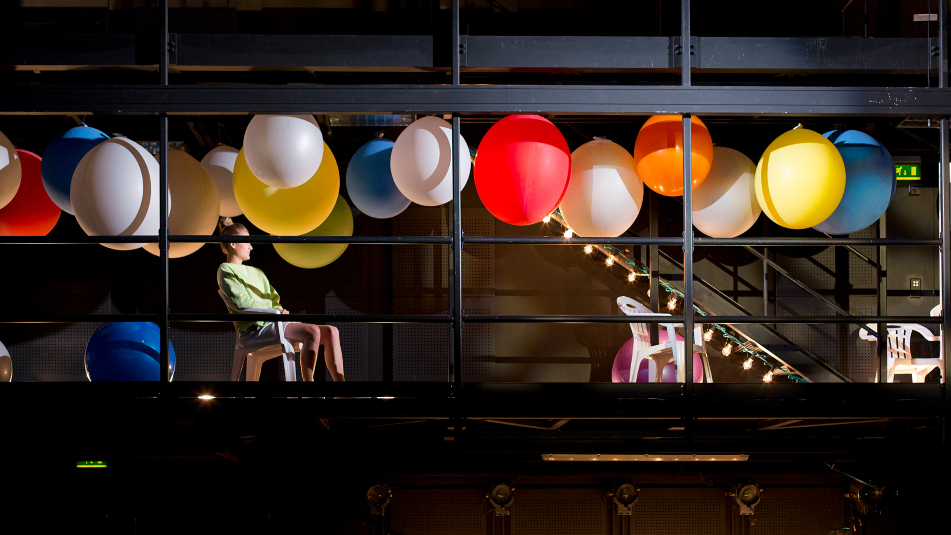 Actor sitting in a chair at a performance stage setting, large colourful balloons above their head.