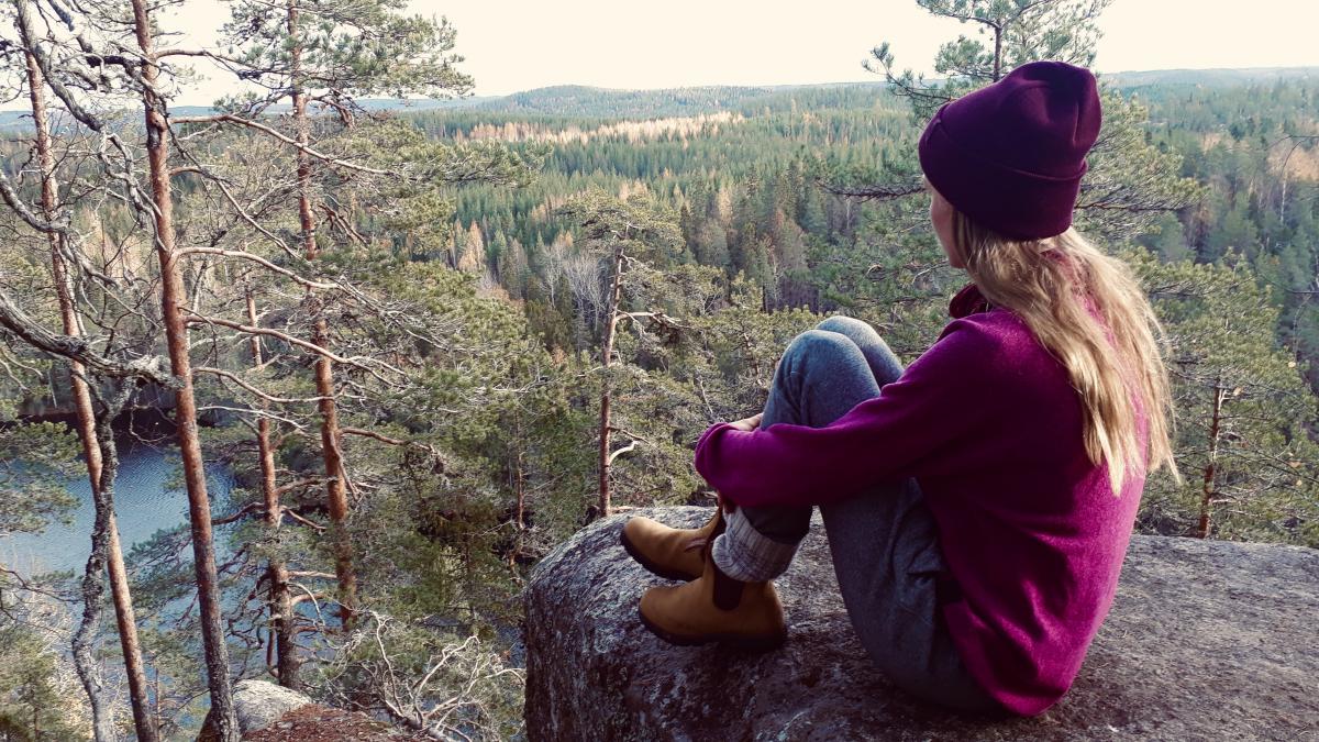 Eerika Grof istuu kalliolla selkä kameraan päin. Taustalla näkyy perinteistä suomalaista kansallismaisemaa, vettä ja metsää.