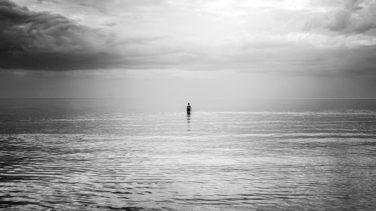 Mustavalkokuva rannalta merelle päin, keskellä kuvaa ihminen seisoo vedessä.
