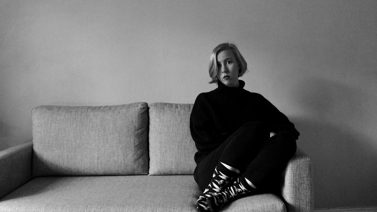 Pinja Savijärvi istuu sohvalla katse kohti kameraa mustavalkoisessa kuvassa.Taustalla on valkoinen seinä, huone on tyhjä. Pinjalla on vakava ilme kasvoillaan.
