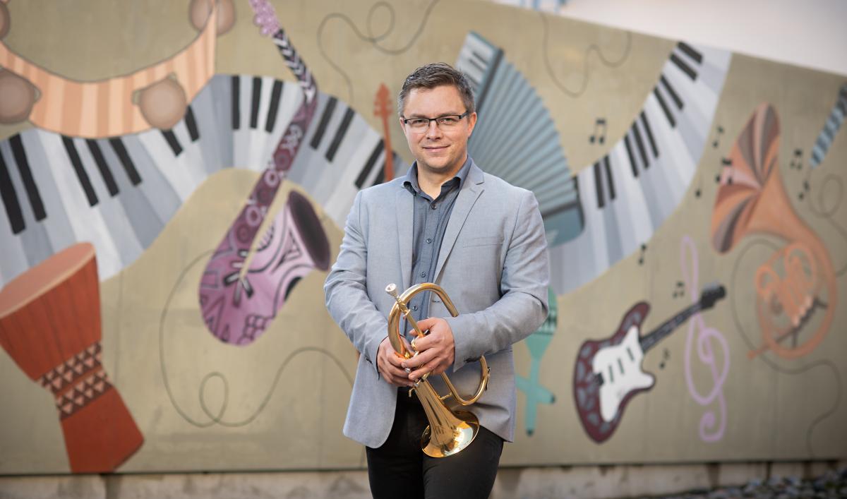 Peter Roos seisoo graffitin edessä, jossa on eri instrumentteja ja hymyilee. Käsissään hänellä on puhallinsoitin.