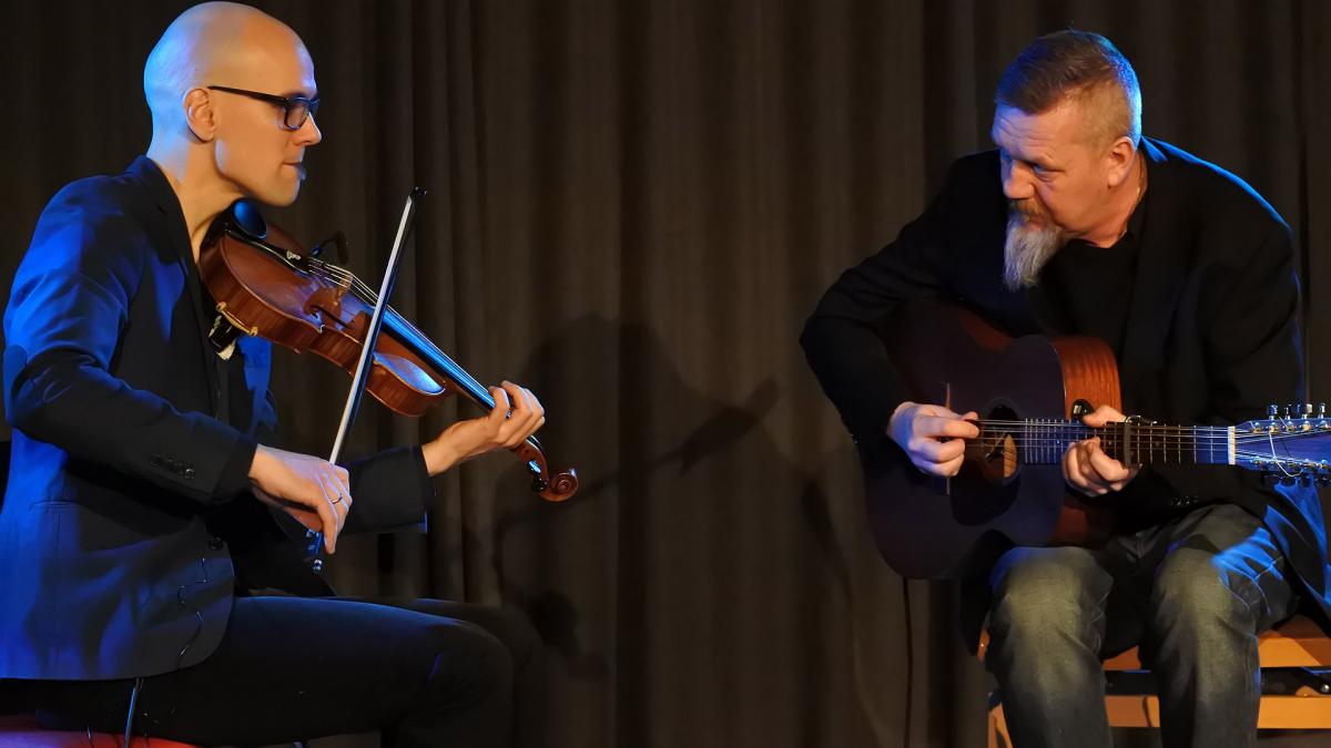 Esko Järvelä & Roger Tallroth soittavat yhdessä. Eskolla on viulu ja Rogerilla kitara. Molemmat istuvat ja tuijottavat toisiaan samalla kun he soittavat.