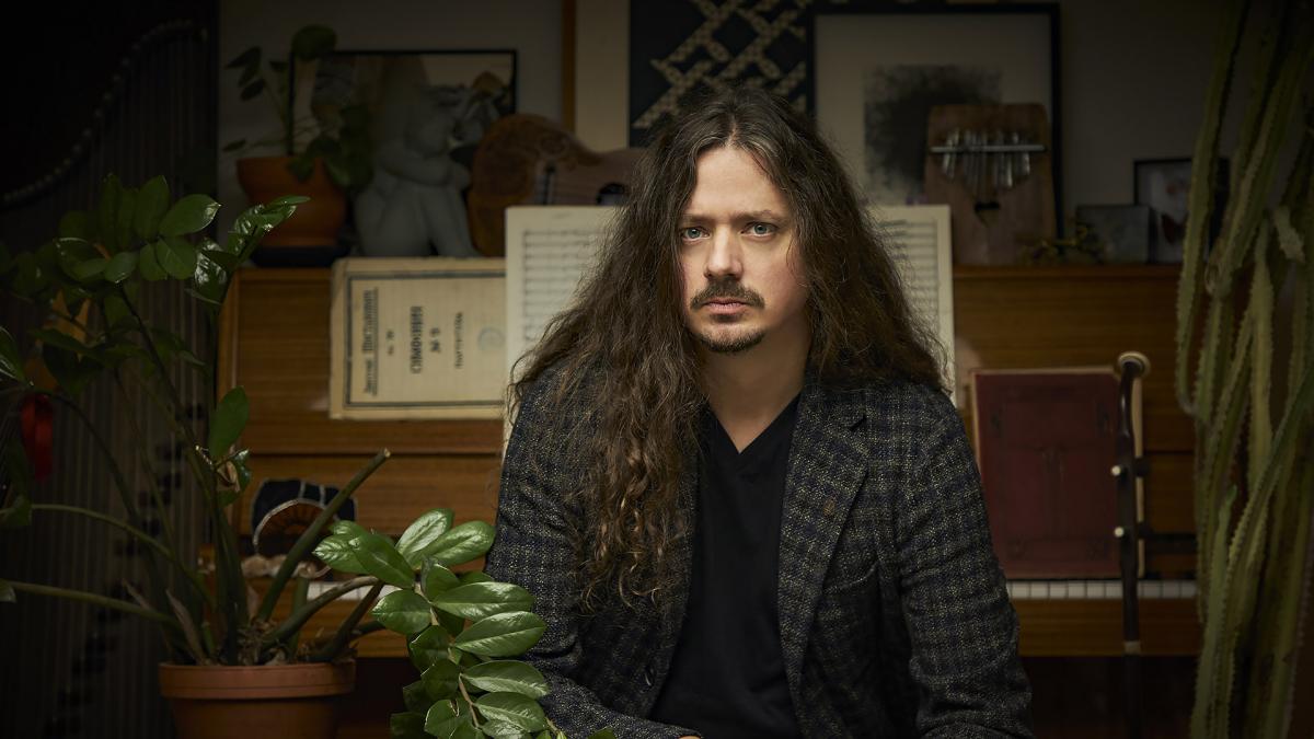 The man is sitting in a room where there are many plants. He looks straight ahead at the camera. The man has quite long hair.