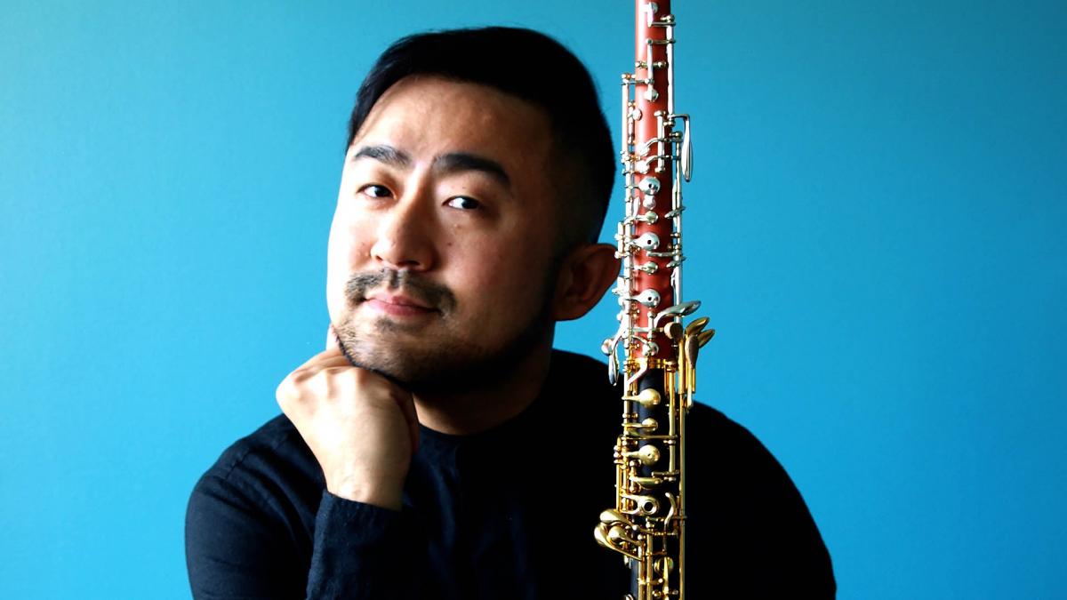 Takuya Takashima leans on his hand and holds oboe with his another hand and smiles gently. The background of the picture is bright blue.