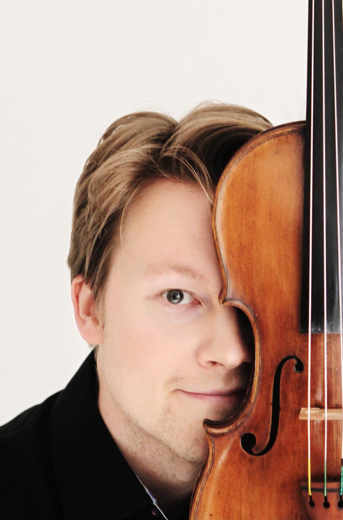 Antti Tikkanen katsoo koti kameraa ja pitää viulua puoliksi kasvojensa edessä. Kuva on otettu studiossa.