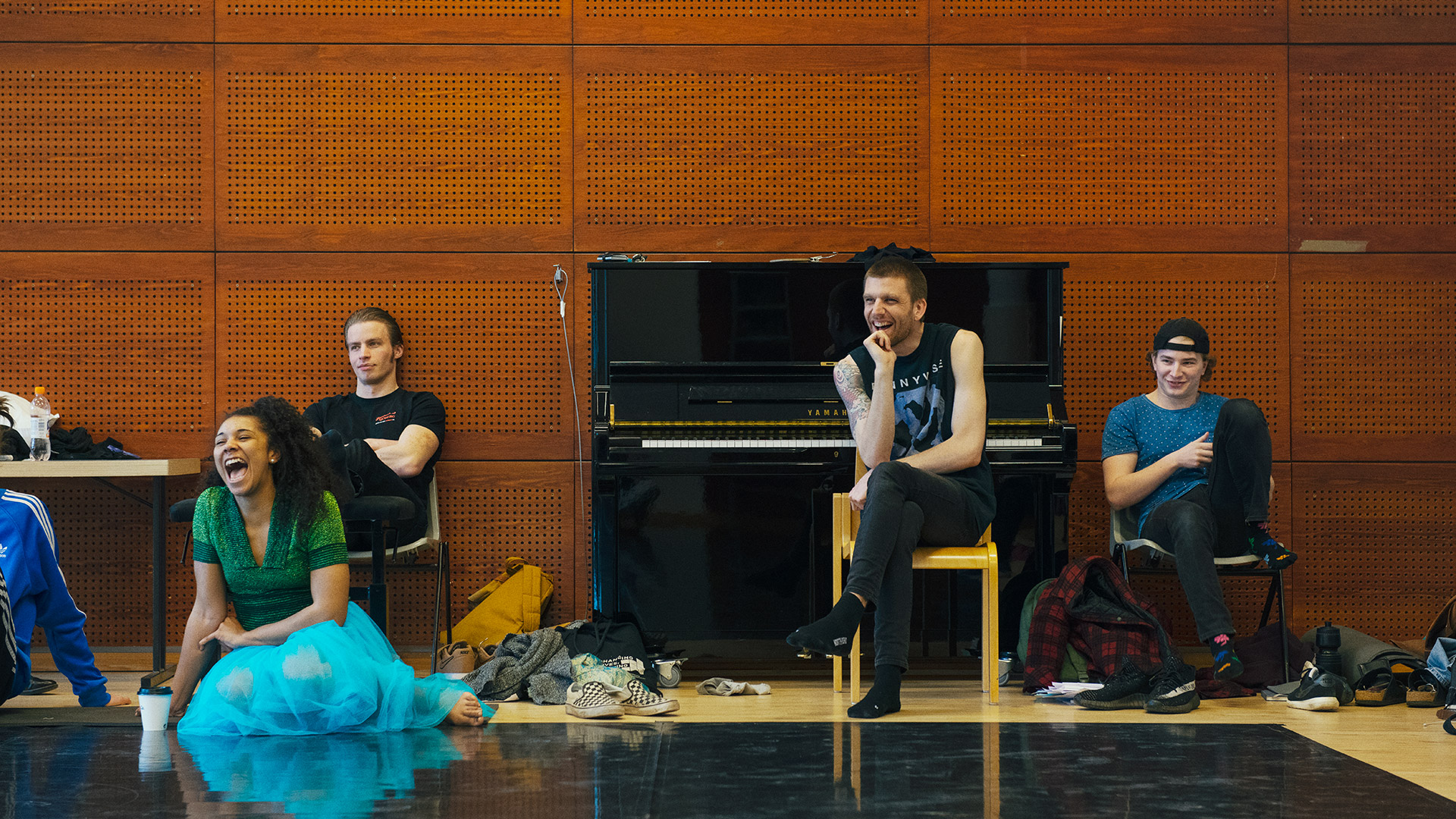 Smalla group of students sitting in a rehearsal space. Some sitting in chairs, one sitting on the floor, laughing. A black piano is in the background.
