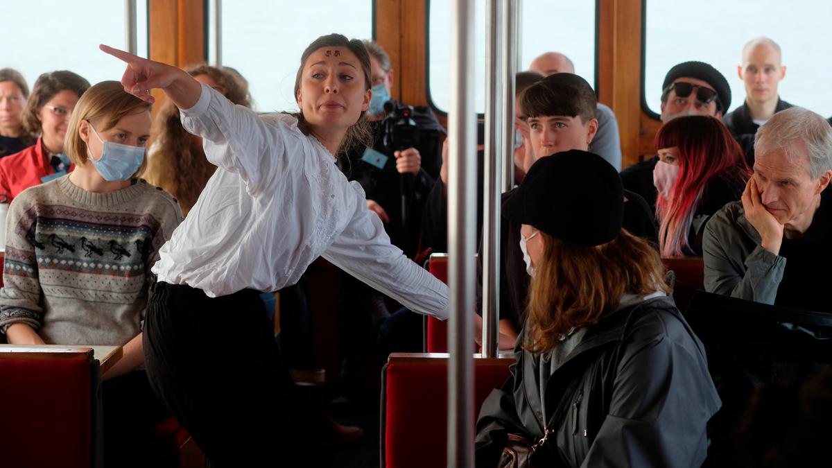 Yleisöä ja esiintyjä lautassa. Esiintyjä osoittaa sormellaan kuvan yläreunaan ja katsoo sitä kohti. Yleisö seuraa esiintyjää istumapaikoiltaan, osalla kasvomaskit päällä
