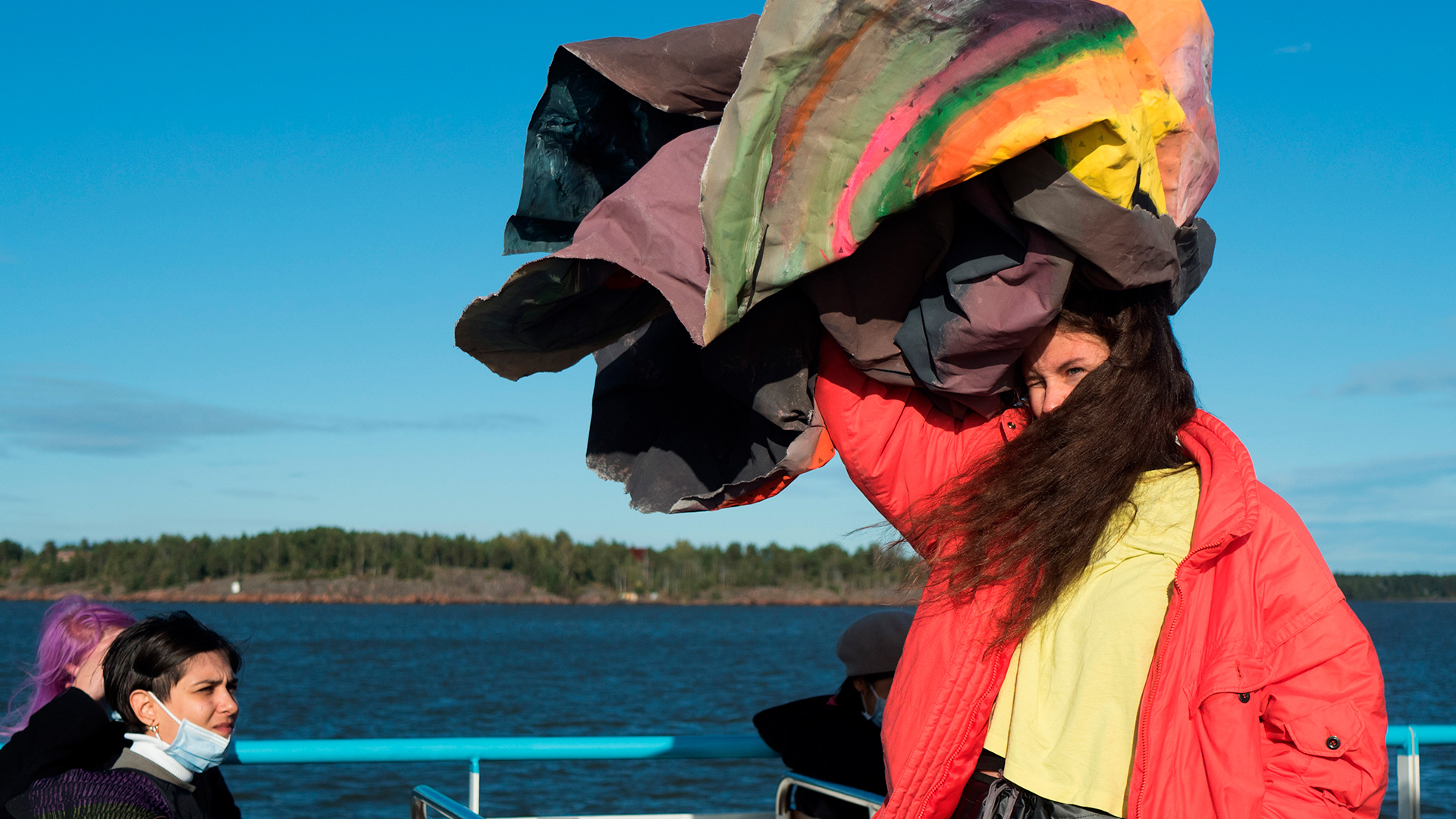 Person with long hair standing outside, where it's windy and sunny. Person is holding a large, colourfully painted, hardened and crinkled canvas above their head. Their hair is partially covering their face.