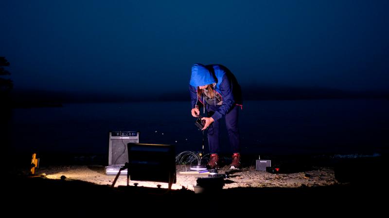 Ulkona pimeässä Dmitri Zherbin tuulipuvussa huppu päässä, hieman kumarassa asennossa, ympärillään ja kädessään audiolaitteita. Maata valaisee yksi valonlähde.