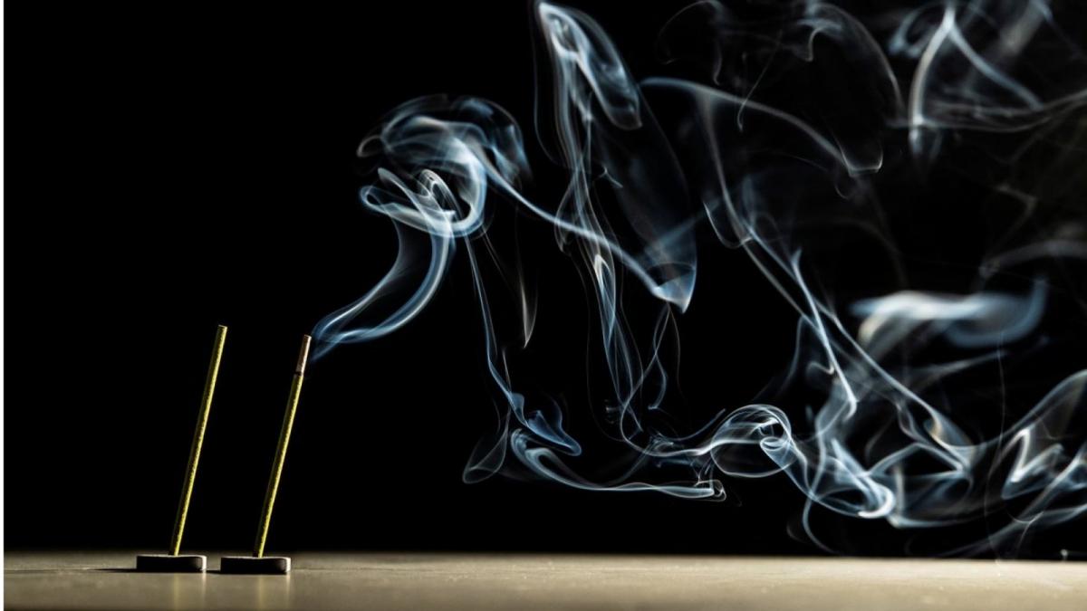 Kaksi suitsuketta palaa pöytäpinnalla ja levittää vaaleana erottuvaa savua ympäröivään tilaan tummasävyisessä kuvassa.