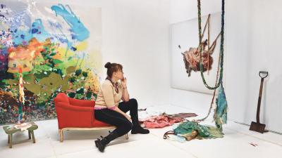 Kuvataiteilija Tarja Pitkänen-Walter katsoo teostaan istuen punaisella divaanilla. Taustalla värikäs maalaus.
