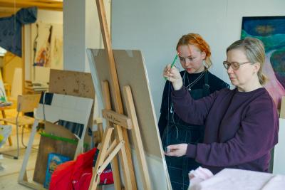 A student and a teacher are standing in front of an easel. The teacher is measuring proportions with a pen while the student observes.