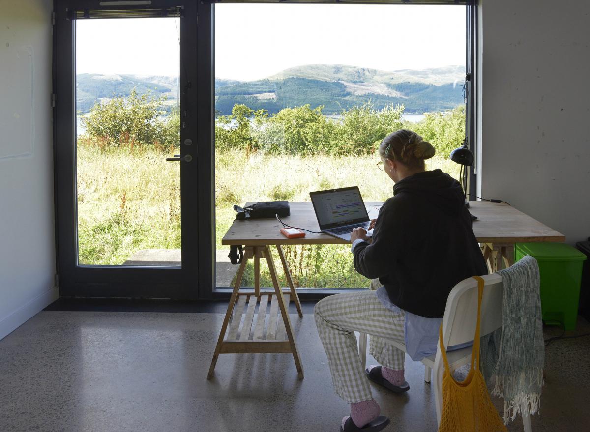 Kuvataiteilija Iisa Lepistö töyskentelee tietokoneella ison ikkunan äärellä Cove Parkissa elokuussa 2022.
