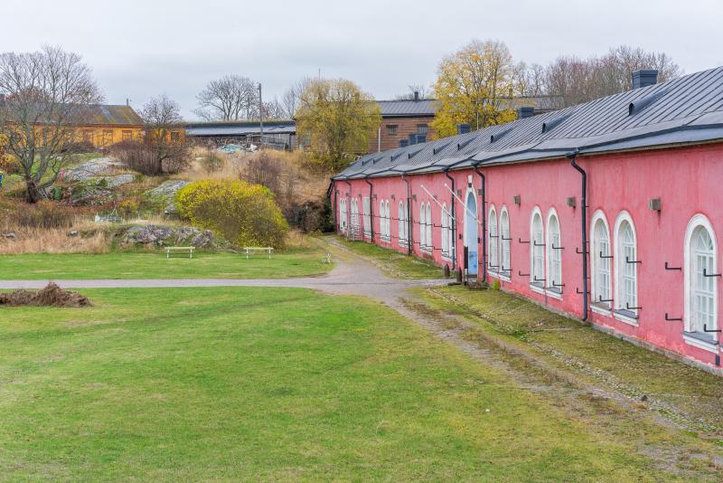 Old pink HIAP gallery fortress building in Suomenlinna at autumn. Green field of grass in front of the building with a walking path and some trees in the background with yellow and orange leaves.