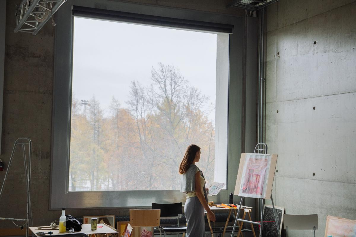Student observing a painting in front of a big window in Mylly building. Through the window a foggy scenery visible.