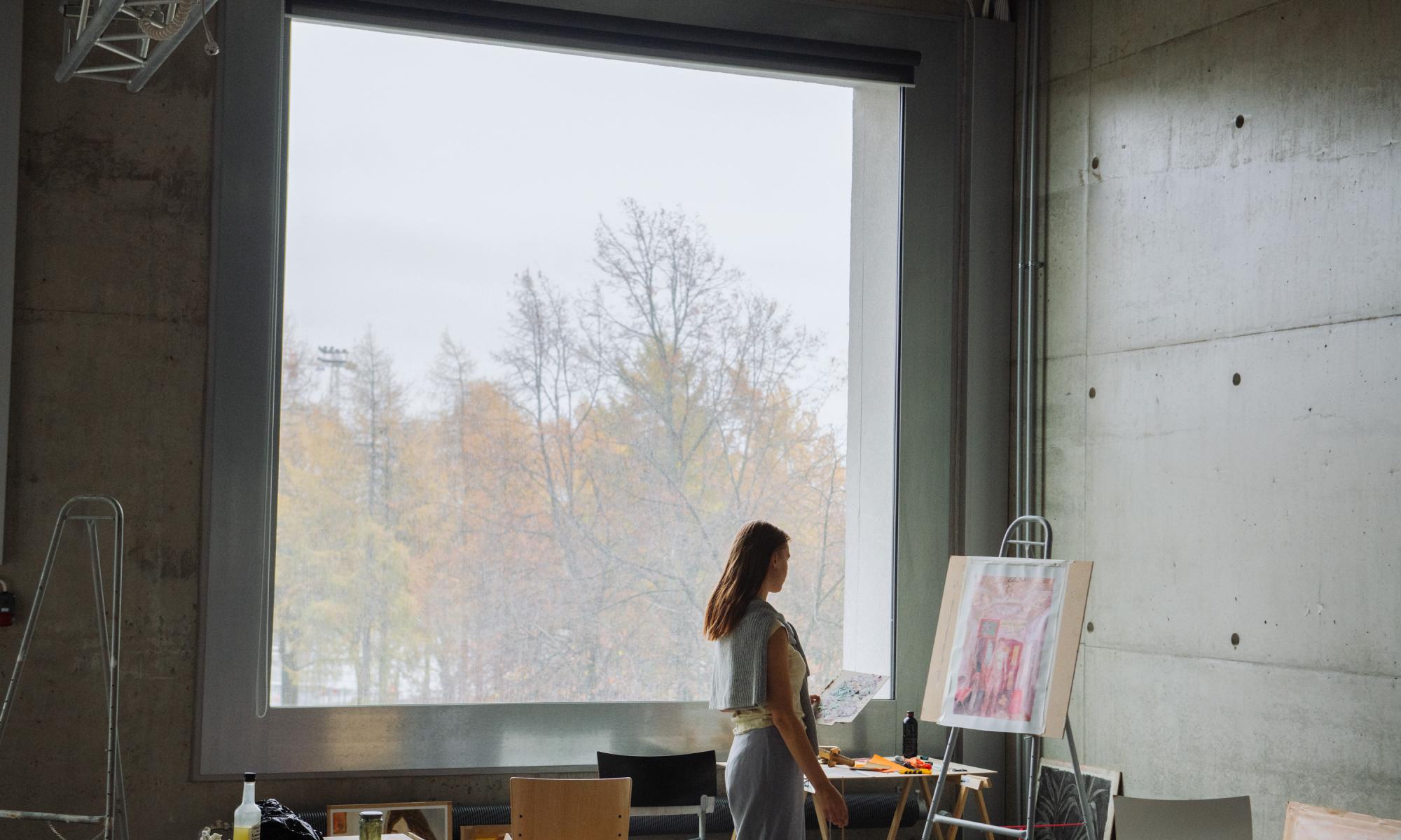Naisoletettu henkilö katsoo keskittyneesti maalausta ison ikkunan edessä Mylly-rakennuksessa, taustalla sumuinen syysmaisema.