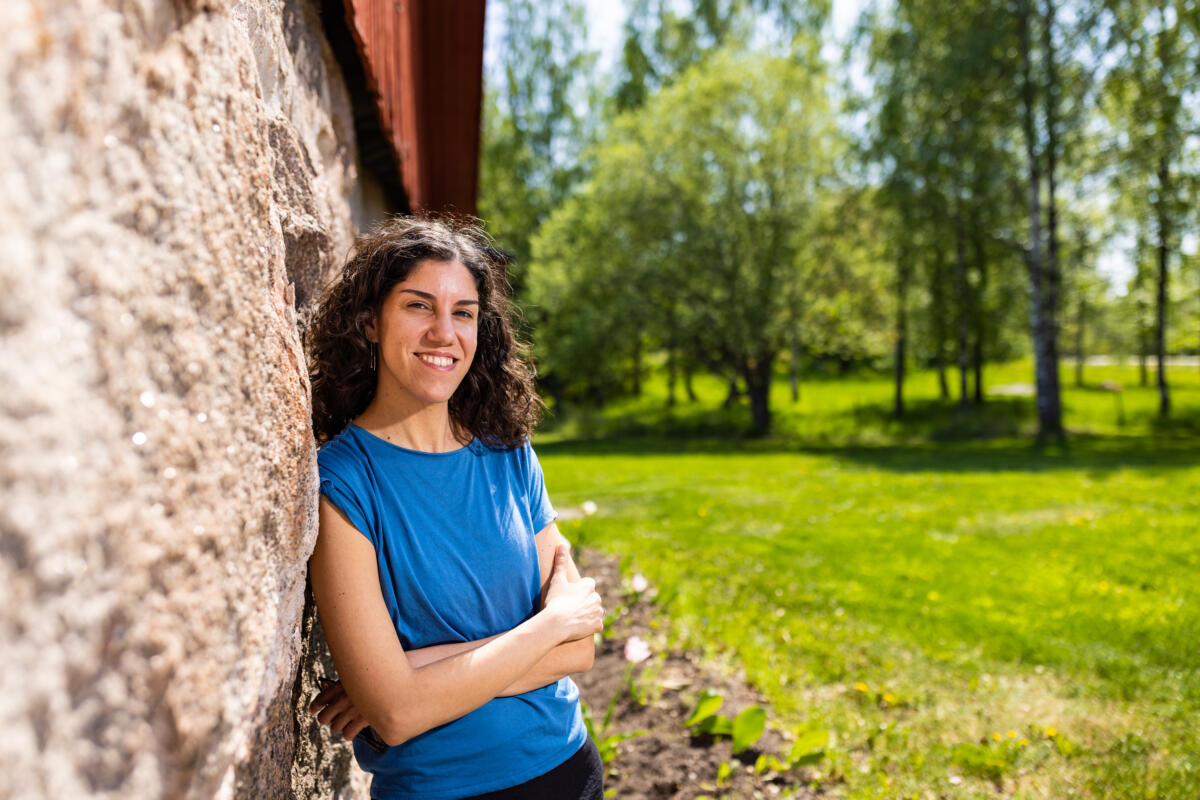Elena Mazzi leans against a stone foundation of an old house smiling at the camera. Behind her there are some trees and green grass.