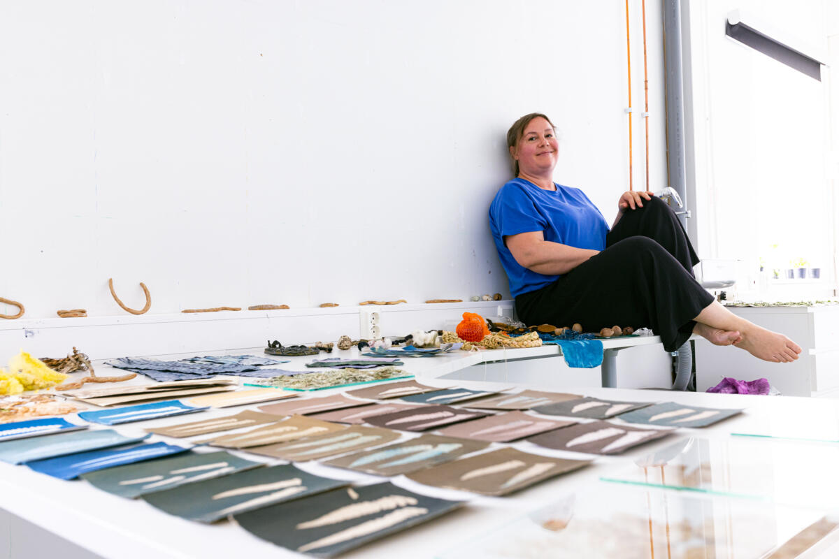 Noora Sandgren sits on a table leaning against a white wall. In front of her on the table there are pictures of leaves in different colours.
